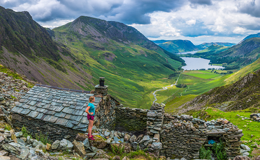 Young female trail runner looking out over a remote mountain bothy towards the picturesque green valley below and the tranquil waters of Buttermere deep in the Lake District National Park, Cumbria, UK.