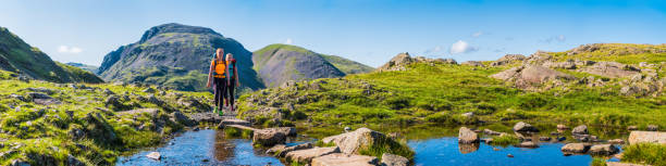 Teenage girl hikers with packs hiking mountain trail wilderness panorama Young female hikers walking along summer mountain trail in the Lake District National Park, Cumbria, UK. english lake district photos stock pictures, royalty-free photos & images