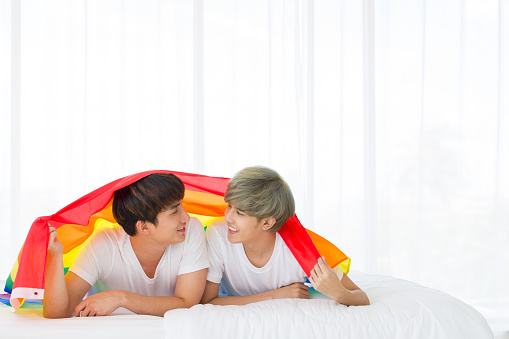 Asian homosexual men or gay couples in a happy time while having a rainbow flag cover body and lying on the white bed. Concept of LGBTQ pride.