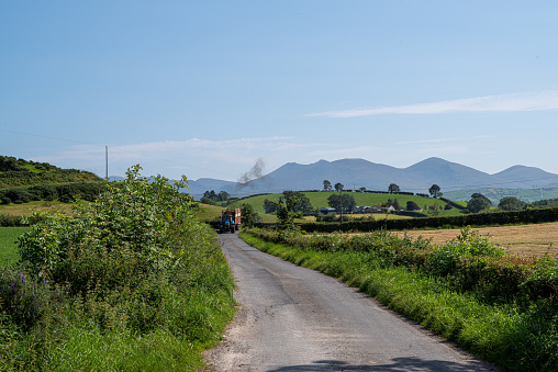 Tractor approaching along a narrow country road, edged by hedgerows.  In the distance is the profile of the Mourne Mountains.  Lackan Bog, County Down, Northern Ireland.