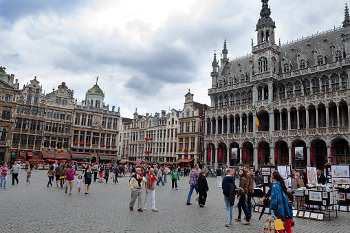 Landmark Grand Place in Brussels in summer at day with overcast sky. Many people and tourists are on square. In right area is Maison du Roi. At right side artists are selling paintings and pictures