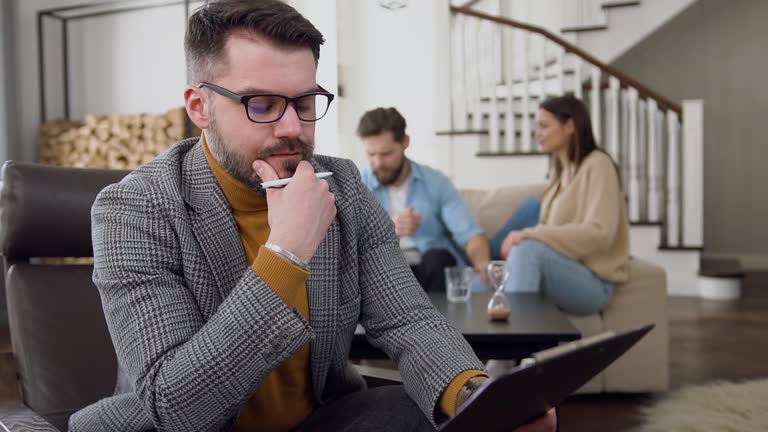 Front view of serious professional bearded psychologist which making notes on the background of smiling young married couple which talking