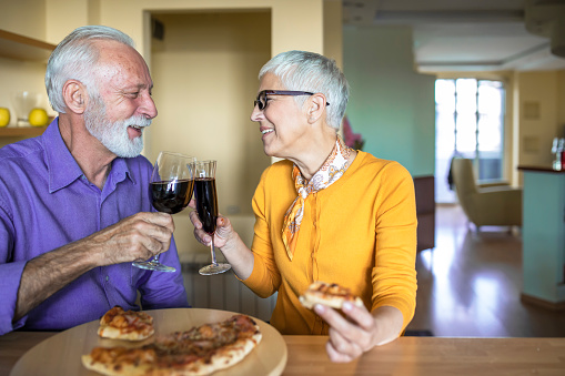 Happy Senior Couple is Sharing Love Emotions while Drinking a Wine at Home and Preparing for Italian Dinner. Senior Man and Woman are Celebrating Anniversary Day While Enjoy in Pizza and Wine.