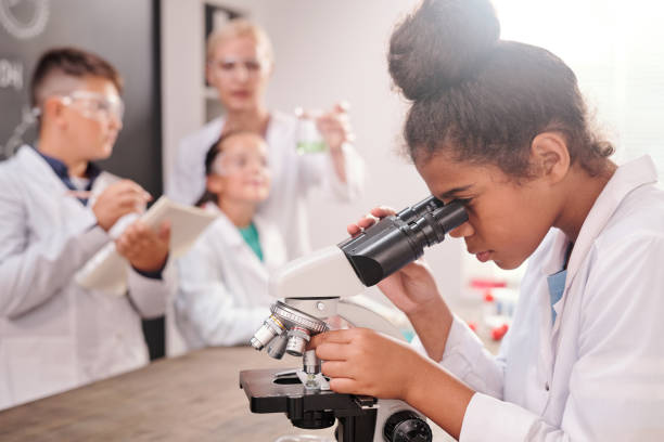 clever mixed-race pupil in whitecoat and eyeglasses looking in microscope - smiling research science and technology clothing imagens e fotografias de stock