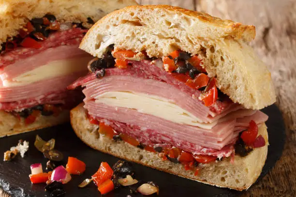 Photo of Authentic New Orleans Muffuletta sandwich filled with olive salad, cheese, salami and ham close-up on a slate board. Horizontal