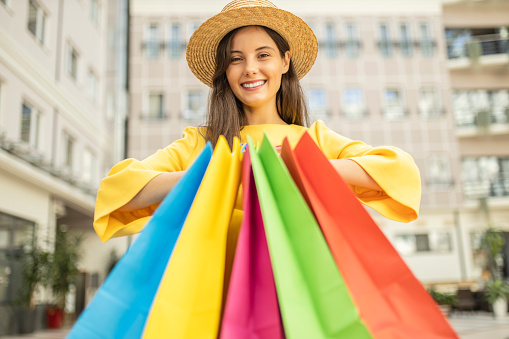 Happy fashionable woman with colorful shopping bags wearing hat and yellow dress and enjoying in shopping mall