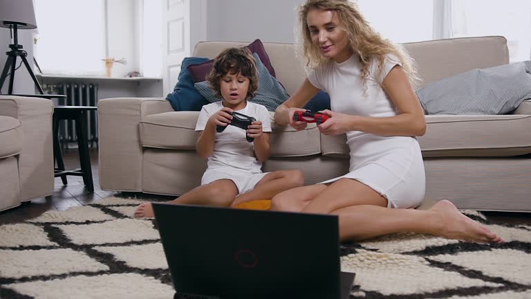 Joyful carefree lovely mother son sitting on the carpet near the couch and playing video game on computer with joysticks