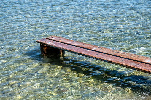 Weathered wooden small pier at Kea, Tzia island, Greece. Wood planks set properly on rusty barrel to make a mini platform over a transparent calm blue sea background.
