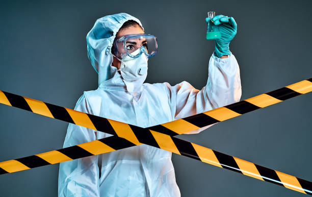 istock Woman in protective suit holding flask with blue fluid behind barrier tape-quarantine 1266607756