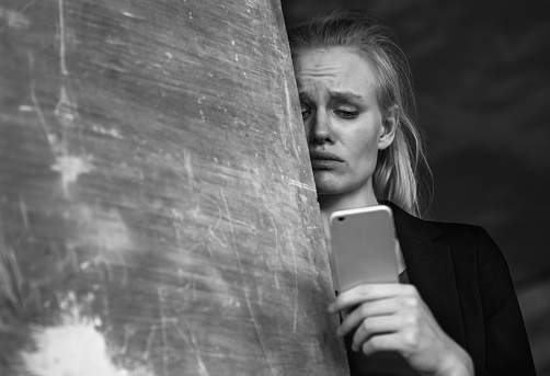 Fear and despair. Depressed caucasian female adult with worried face expression receiving bad news on her smartphone. Black and white closeup portrait.