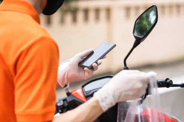 Courier deliveryman with face mask and glove on motorbike or motorcycle check customer location by smartphone. New normal of food delivery business during covid19 or coronavirus pandemic concept.