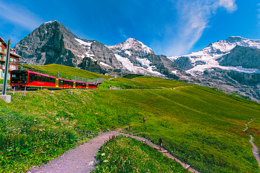 Kleine Scheidegg, Bernese Oberland, Switzerland - August 1 2019 : Famous red train to Jungfraujoch with snow capped Eiger north face, Monch and Jungfrau in the background