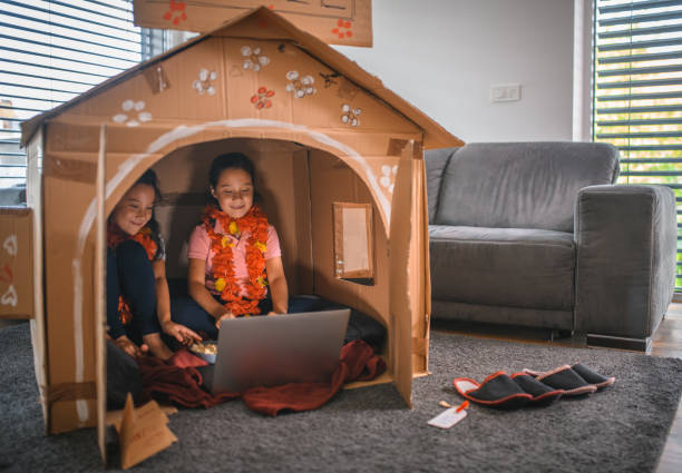 Asian sisters watching movie in cardboard house during quarantine time Asian sisters are having movie night in cardboard house at home in a living room. They are wearing Hawaiian necklace. They are spending fun time together during quarantine time. playhouse stock pictures, royalty-free photos & images