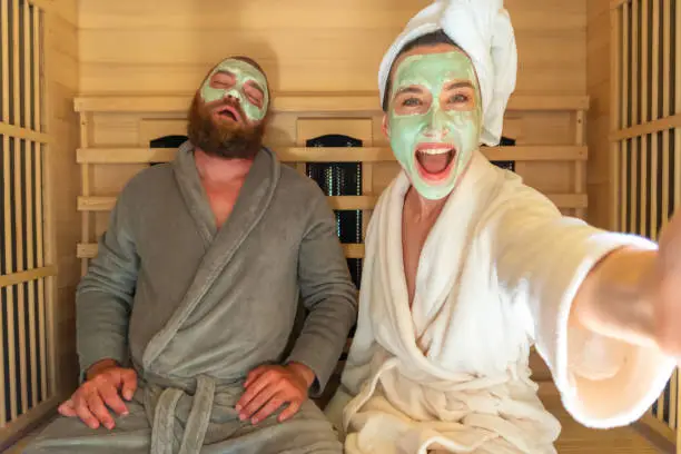 Photo of A young funny couple having relax SPA day in sauna, a woman making selfie with beauty mask while her boyfriend is asleep.