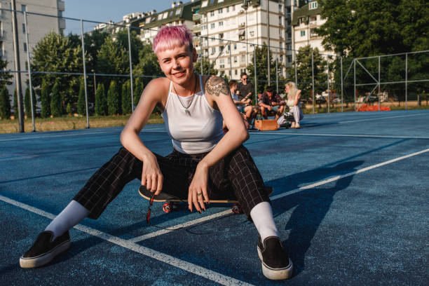 skateboard-mädchen mit rosa haaren sitzen vor einer gruppe von freunden auf einem sportplatz - skateboard court stock-fotos und bilder