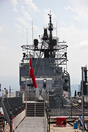 The forward turret and 5 inch Mk45 Mod 2 automatic rapid fire gun of HMAS Arunta, an Anzac Class frigate of the Royal Australian Navy.  She is docked at Garden Island, Sydney Harbour. In the background is a pole with six visible security cameras. This image was taken on a hot and sunny afternoon from Woolloomooloo Bay on 3 March 2024.