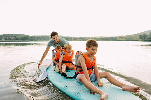 Photo of a young father and his sons getting ready for the paddleboard ride on the lake; having hun and enjoying the beautiful summer afternoon.