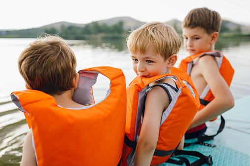Photo of three little boys sitting on the paddleboard, taking a short break after swimming; spending time outdoors on a hot summer afternoon.