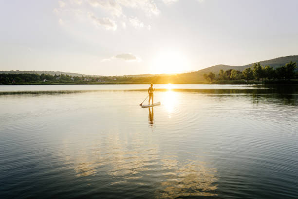 A young adult man floats on his standup paddleboard in the salt water of the Puget Sound, an inlet of the Pacific Ocean in Washington state, in the United States.  The sun casts pink and orange colors onto the water.