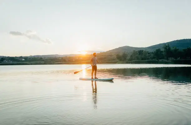 Photo of a young man stand-up paddleboarding on the lake; enjoying the beautiful, warm summer afternoon, far from the hustle of the city.