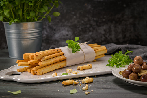 Fresh baked homemade grissini bread sticks in white ceramic tray with olive oil and basil herbs over kitchen counter top.