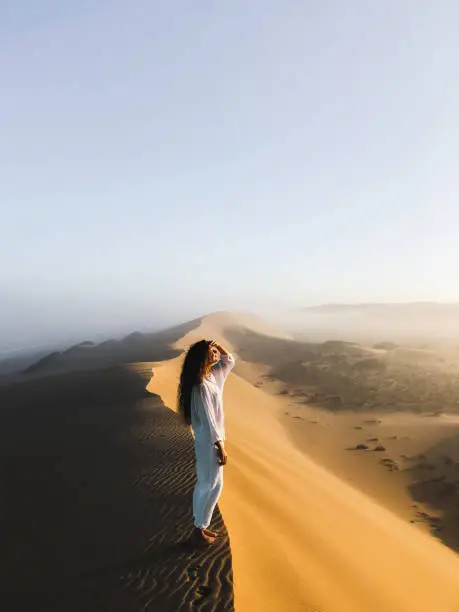 Photo of Woman enjoying sunrise on top of huge sand dune. Beautiful warm sun light and mist in morning. Sahara desert, Morocco.