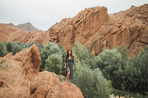 Portrait of beautiful european woman barefoot on background of Todra gorge canyon in Morocco. Long black dress and tattoo on naked leg.
