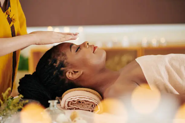 Therapist holding hands over head of pretty young Black woman to transfer energy during healing reiki session