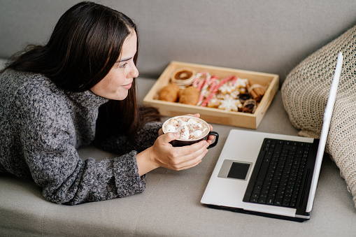 Cozy winter weekend. Holidays atmosphere, warming, hygge concept. Woman in sweater enjoy traditional sweets and watching tv series on laptop in her living room. Leisure and entertainment