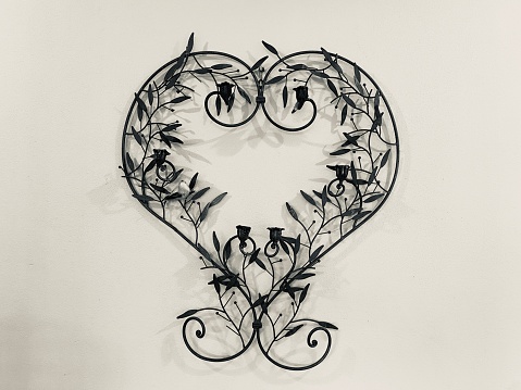 Horizontal photo of a black metal heart-shaped wall candleholder on a wall. Leaves and buds decorate the candleholder.