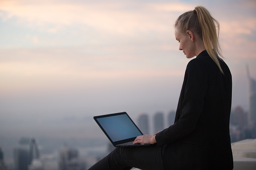 Relaxed female business person peacefully busy on a laptop computer.  She is working alone sitting on a skyscraper rooftop with a beautiful city view during sunset.