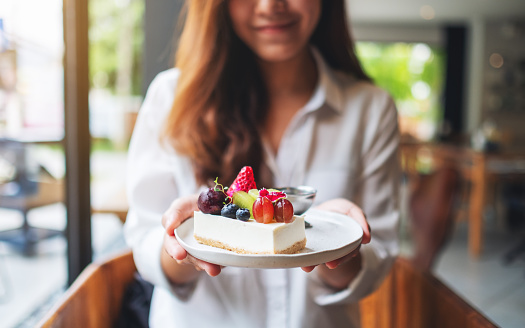 A beautiful young woman holding and showing a plate of mixed fruits cheesecake in cafe