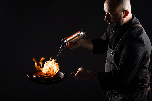 Chef cooks sausage, roasts meatballs on open fire. Black background.