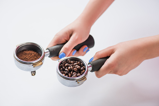 cropped view of barista holding portafilters with coffee beans and ground coffee on white background