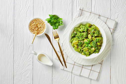 Italian gnocchi with broccoli tossed with basil pesto and creamy cheese sauce sprinkled with pine nuts in a white bowl on a wooden table, close-up, horizontal view from above, flat lay