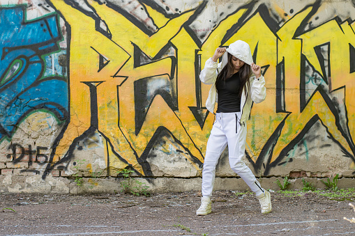 Young girl in a white tracksuit dancing against the background of a wall with yellow graffiti