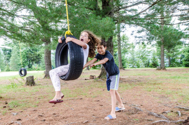 Boy helping sister in a swing Boy helping sister  in a swing made with a car tire attached to a tree during summer day. tire swing stock pictures, royalty-free photos & images