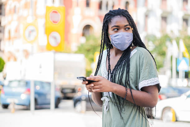 istock Young woman wearing protective face mask walking through city, using smart phone 1266759358