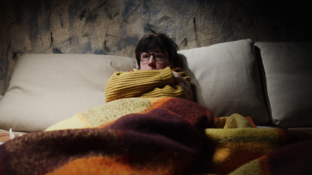 Mature woman is wrapping herself with shawl and blanket while getting fever on the sofa