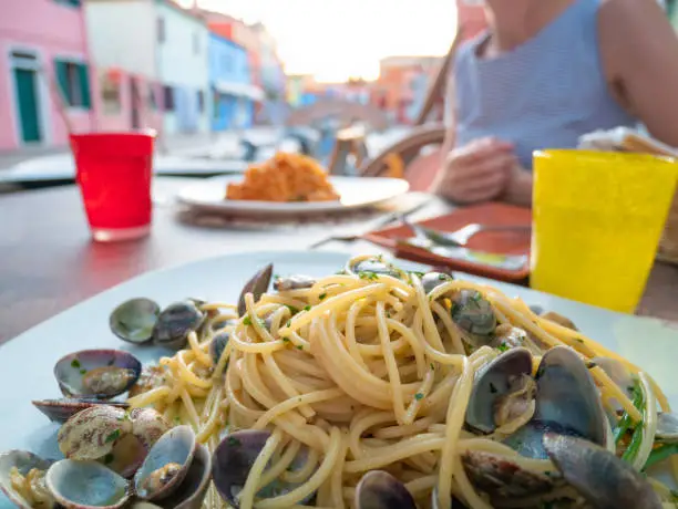 Romantic dinner for two in colorful town Burano, Venice, Italy. Italian Pasta Spaghetti with clams and spaghetti with seafood, Beautiful woman eats at a Restaurant Table along the canal near the boat