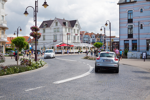 Traffic on s-shaped curved street in De Panne in summer. People are walking on sidewalk. In street are old buildings. De Panne is popular town at North Sea coast