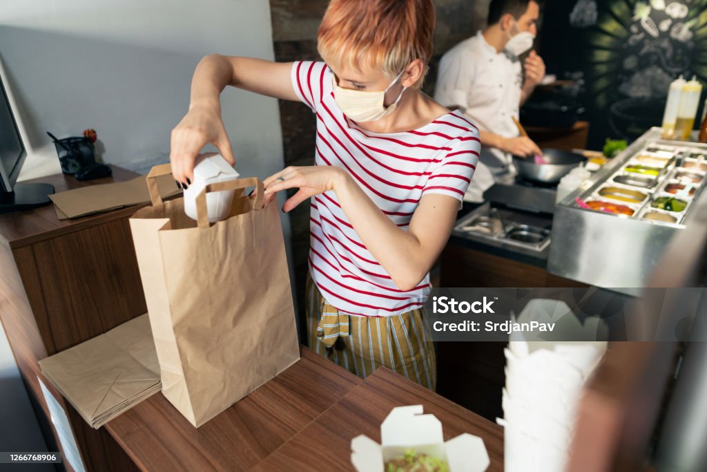 Food workers Fast food restaurant during coronavirus pandemic. Female fast food worker with protective face mask packing meals for delivery while her male coworker preparing food in the back. Delivering Stock Photo