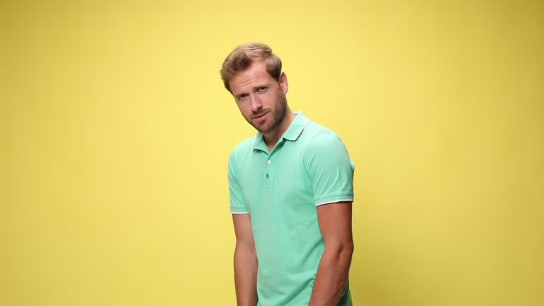 timid young casual man holding hands on hips and standing in a side view pose on yellow background