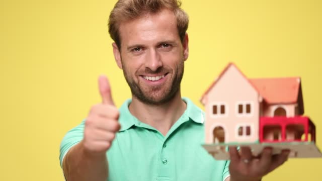 happy casual man holding and recommending house, making thumbs up and smiling on yellow background
