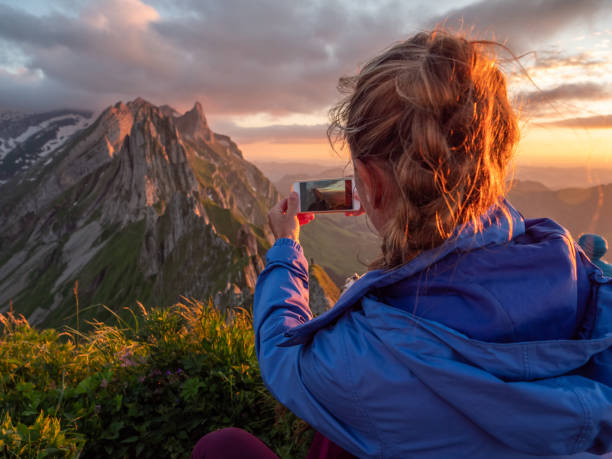 istock Woman photographing sunset from mountain peak using mobile phone 1266778782