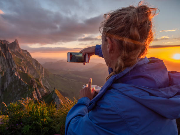 istock Woman photographing sunset from mountain peak using mobile phone 1266780889