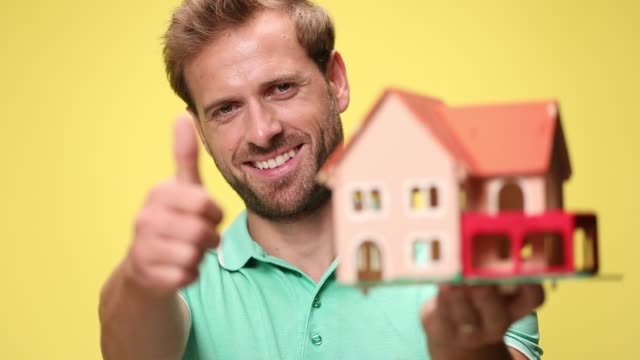 young man admiring house, holding and presenting house, making thumbs up gesture and smiling on yellow background