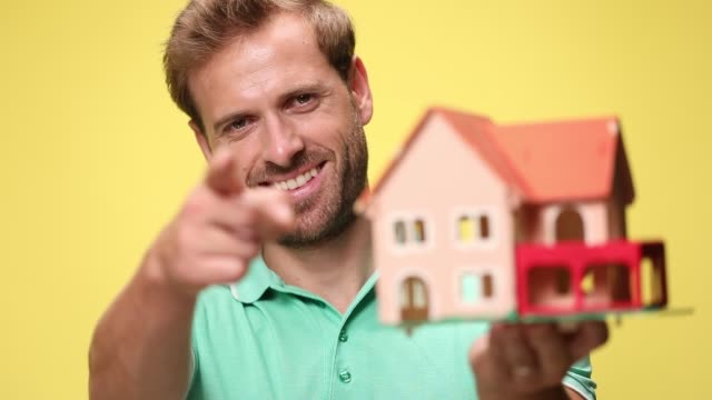 smiling young casual guy holding up house model, recommending and pointing fingers on yellow background