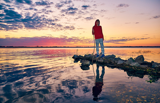 A teenager in a red hoodie and gray pants and blue sneakers stands on the stone pier of the river. The boy holds a stick in his hand. Raspberry sunset on the shore. Mirror reflection of a cloudy sky in the water.
