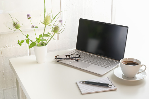 White office desk with laptop, glasses, notepad, flower bouquet and a coffee cup, small workplace at home or in a modern company against a bright wall, copy space, selected focus, narrow depth of field